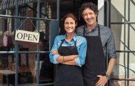 Two cheerful small business owners smiling and looking at camera while standing at entrance door. Happy mature man and mid woman at entrance of newly opened restaurant with open sign board.